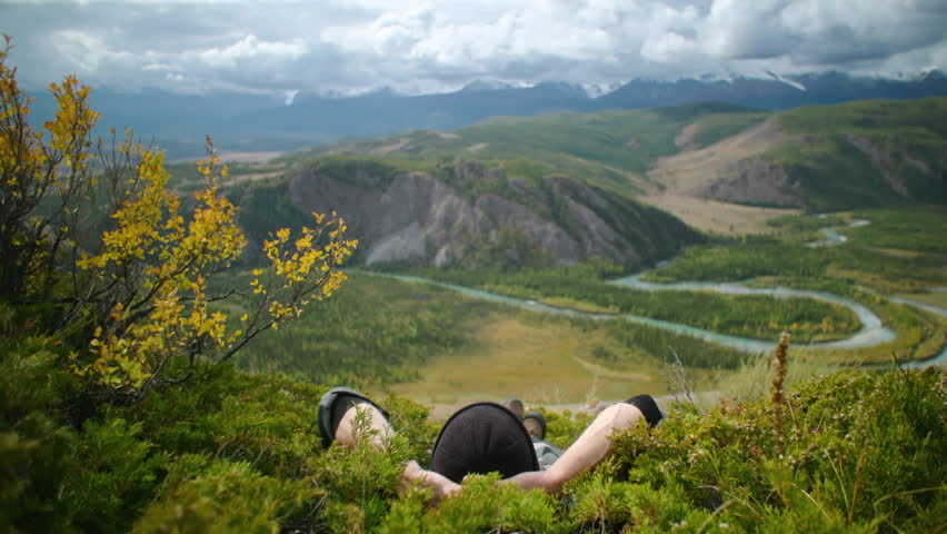 Handsome Man is lying on the grass in the background view of majestic mountains and a winding river, inviting exploration of adventure and natural beauty
