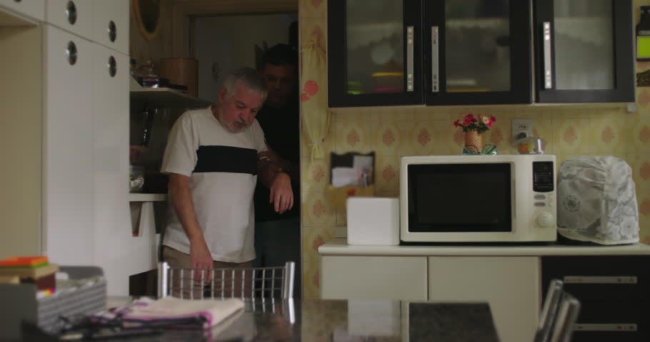 Male Caretaker helping an elderly man with dementia or age-related illness from the bedroom to sit down in the kitchen chair, providing care and support with compassion
