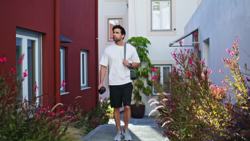 Sporty man walking outdoors with fitness gear surrounded by flowers greenery. Young sportsman going training carrying sport equipment on street. Unshaved guy athlete enjoying active healthy lifestyle.
