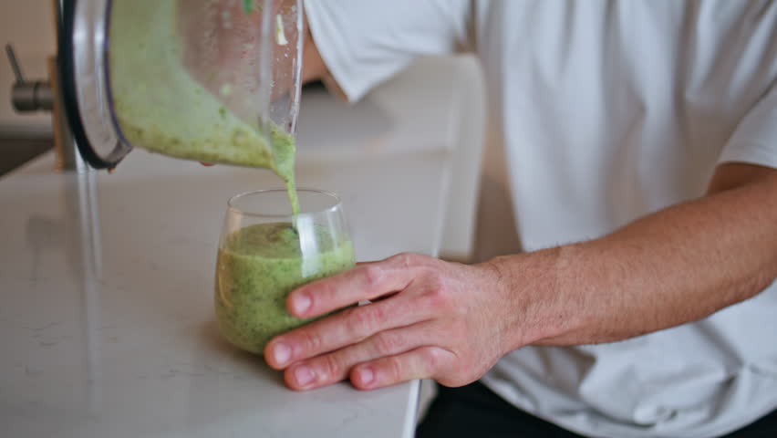 Hands pouring fresh smoothie from blender to glass on white kitchen countertop closeup. Unknown man make healthy homemade beverage for body wellness. Diet sporty nutrition preparation in apartment.