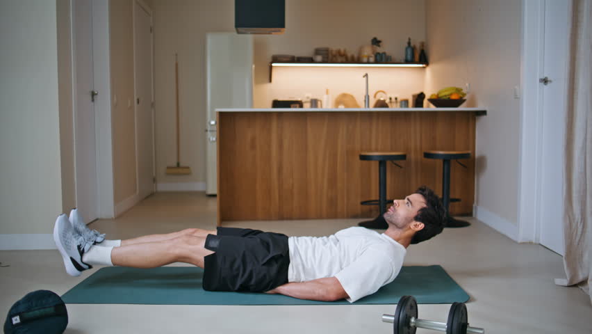 Fitness guy raising legs lying on sport mat at core training exercises. Determined sportsman pumping abdominal muscles on apartment floor. Active hispanic man training muscular healthy body indoors.