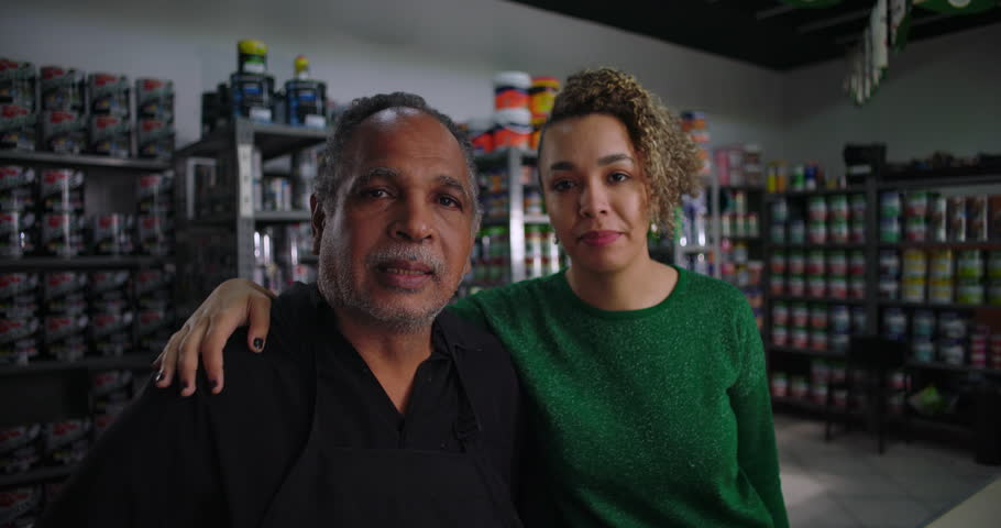 Father and daughter standing close together in their family owned hardware store, shelves of paint cans and supplies in the background, showing teamwork and family bond