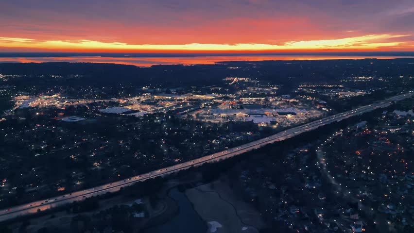 As the view unfolds it presents the consistent evolution of commuters across a highway as they pass an area of plazas and shopping centers underneath the glow of the sunset and the reflection of hues 