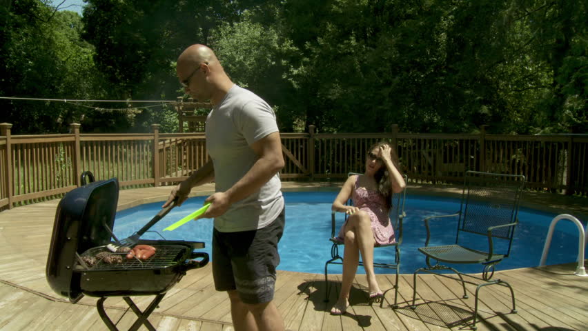 Couple sat in chairs at poolside. Man gets up and serves up BBQ from grill.