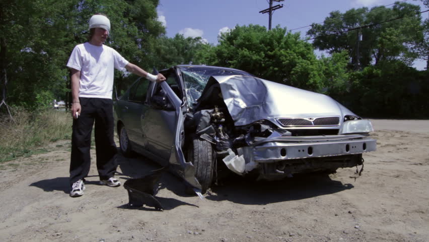 Bandaged teenage driver stands by the wreckage of his car and looks at his