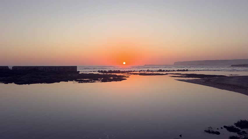 The sun is setting over the Atlantic Ocean in Barbate, Andalusia, Spain, casting a warm orange glow on the water and the rocky shoreline, creating a peaceful and serene atmosphere