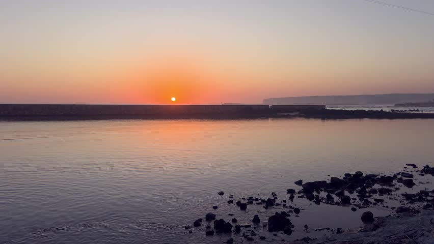 Sunset casting a warm orange glow over the horizon in Barbate, Andalusia, Spain, reflecting beautifully on the calm water with rocks and a breakwater in the foreground