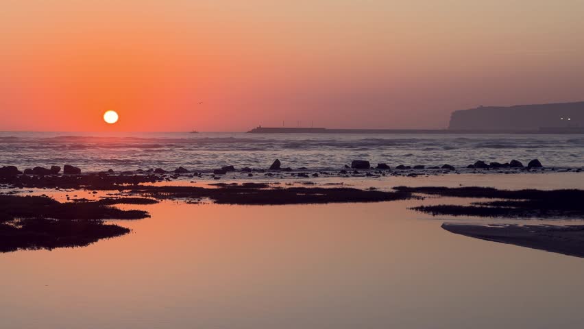 The orange sun sets over Barbate, Andalusia, Spain, casting a warm glow on the wet sand and rocks at low tide, with a breakwater and cliff in the distance