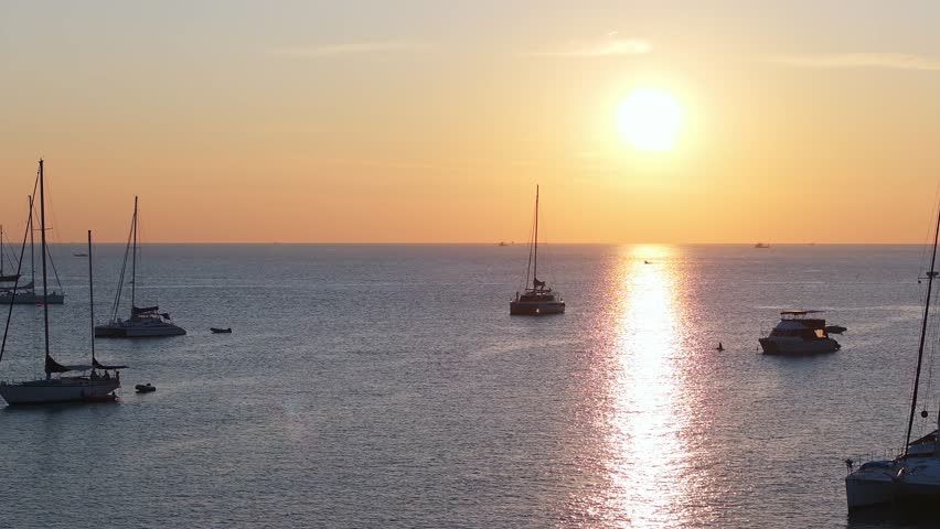 Serene sunset over calm ocean near Phuket Island, Thailand, with sailboats and yachts floating. Golden sunlight reflects on the tranquil water.