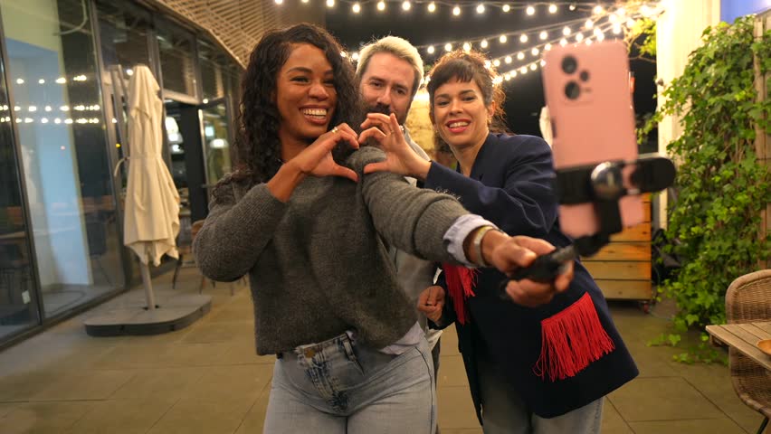 Multi-ethnic friends join hands gesturing heart shape while vlogging online in a night party