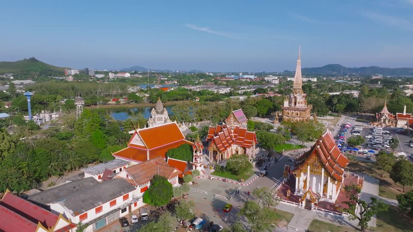 Aerial view of Wat Chalong temple in Phuket, Thailand, featuring its golden spire, lush greenery, a parking area, a market, a lake, and distant hills.
