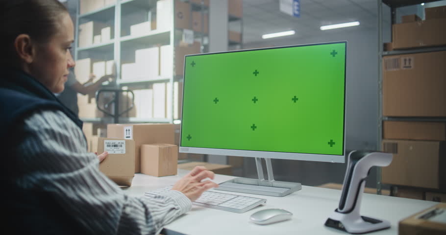Female Warehouse Logistics Manager Scans Code on Cardboard Box with Scanner, Works on PC in Logistics Center, Prepares Package for Delivery. Computer Screen Shows Green Screen Chroma Key. Handheld.