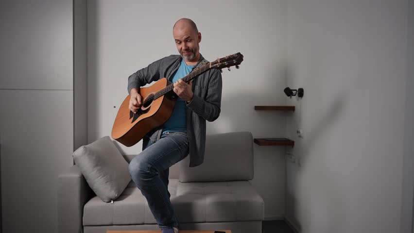 A man passionately plays an acoustic guitar while standing in a simple room. His facial expression reflects the emotional intensity of his music, with a strap securing the guitar around his shoulder.