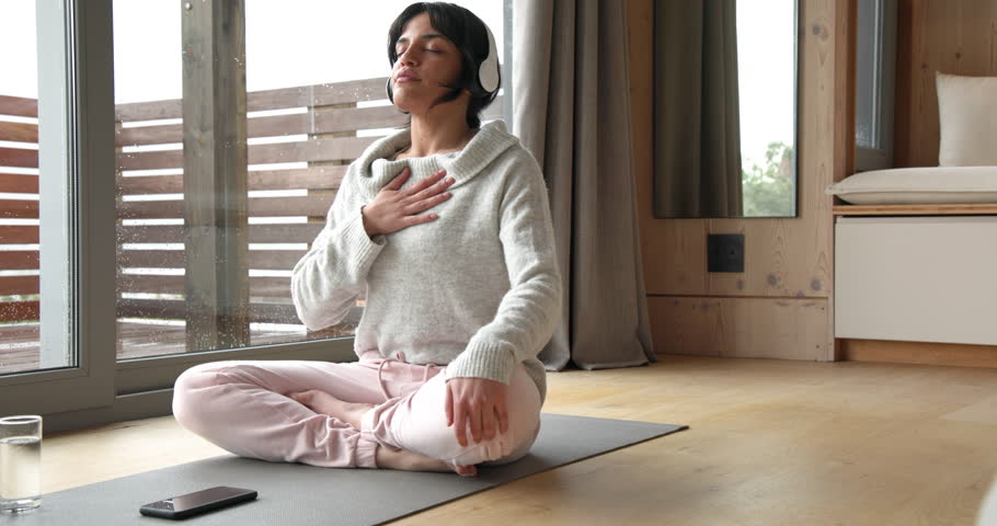 Woman meditating on yoga mat at home, wearing headphones, feeling relaxed. Meditation, relaxation, wellness, mindfulness, peaceful, zen