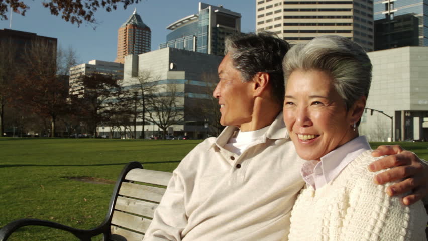 Older Asian-American man and woman sitting on a park bench with the city of Portland, Oregon in the background. Medium close up with dolly movement.