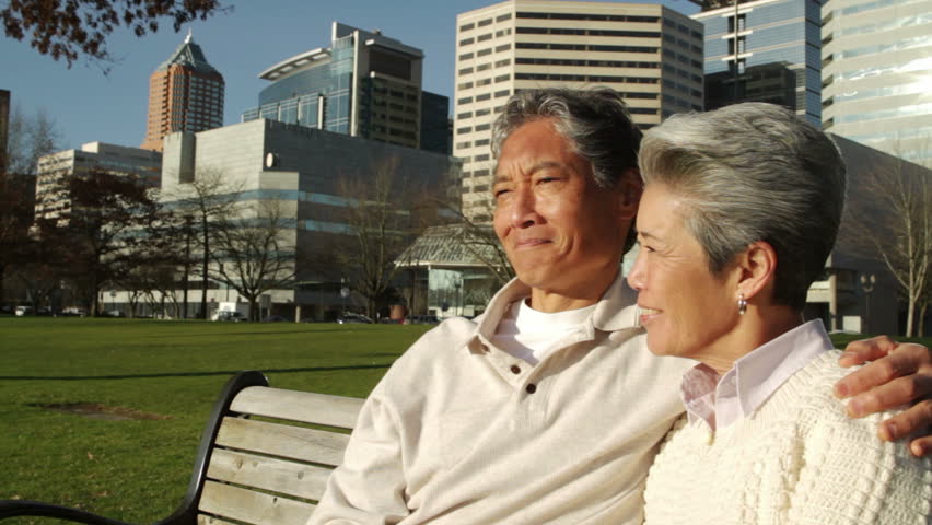 Older Asian-American man and woman sitting on a park bench with the city of
