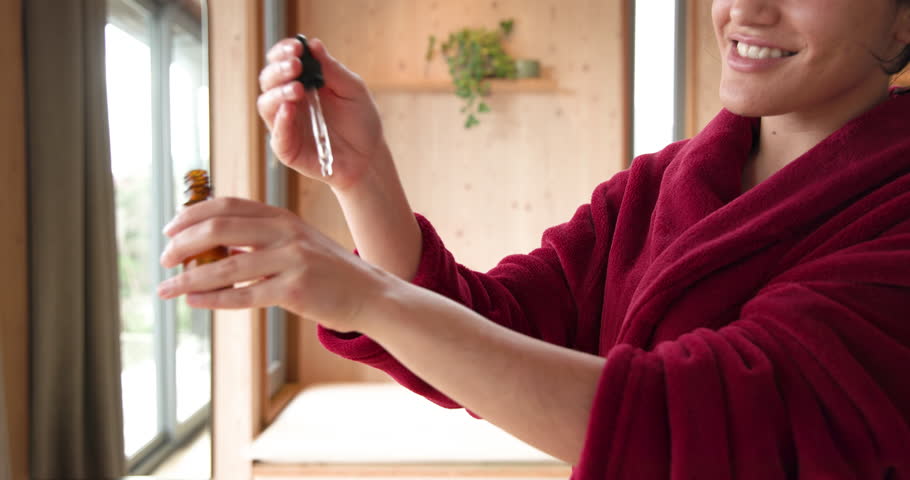 Applying essential oil on hands in cozy home wearing red robe. Aromatherapy, relaxation, wellness, skincare, beauty, self-care