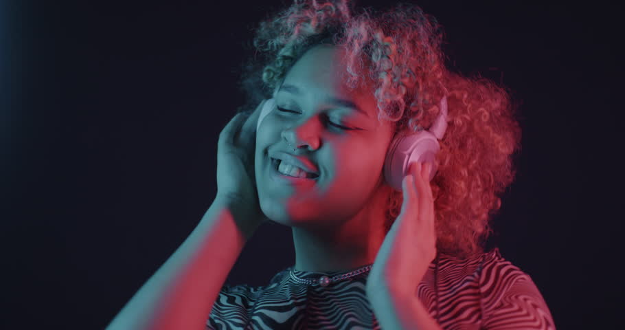 African American woman listening to music with headphones dancing in neon light on dark background. Entertainment and people concept.