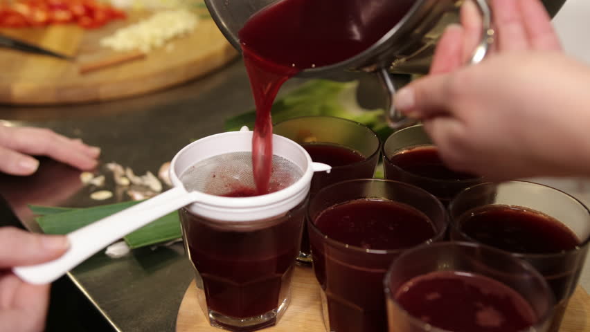 Kitchen counter, fresh ingredients, healthy drink. Close-up shows hands pouring dark red juice through strainer into glasses, with vegetable scraps scattered on kitchen counter.