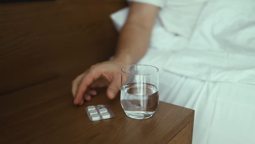 Closeup of young Caucasian man sitting on bed with glass of water, taking sleeping pills or painkillers at home. Unrecognizable millennial male using medications, holding tablet in hand, selective
