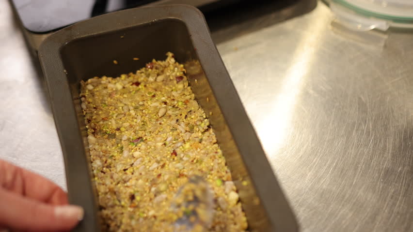 Seeds mixture, spoon mixture, homemade dish. In close-up view, person uses spoon to press mixture seeds, nuts, and grains into rectangular baking tin.