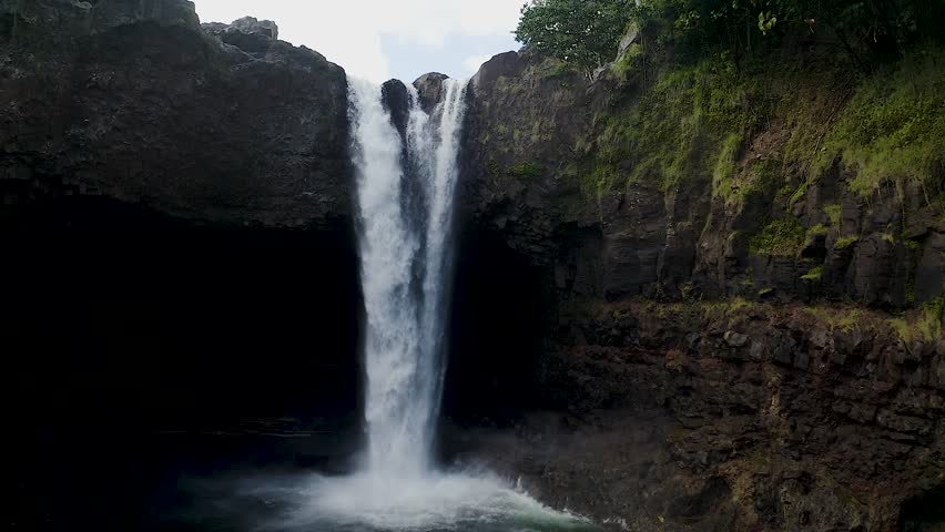 A stunning slow-motion close-up of Rainbow Falls on Big Island, Hawaii, with powerful cascading water flowing into a rocky pool below.