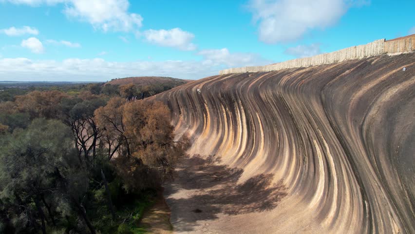 Aerial view of wave rock and surrounding trees under a blue sky with clouds, Hyden, Australia.