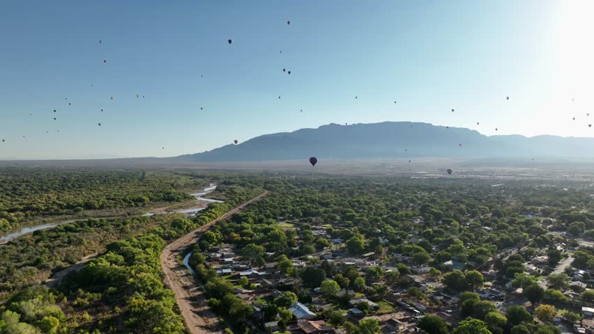 Drone POV of multiple hot air balloons flying over Albuquerque, New Mexico in the early morning near the Sandia Mountains.
