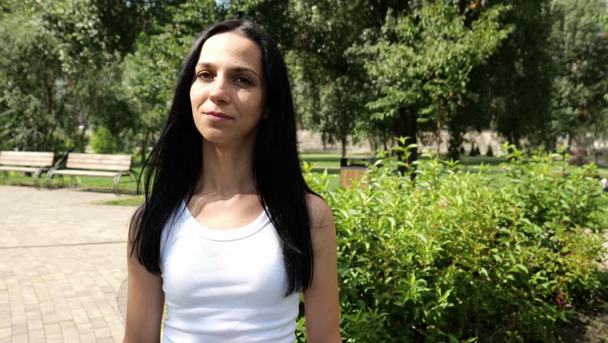 Portrait of a young brunette woman in a white T-shirt walking along the road in the city park. Slow motion portrait