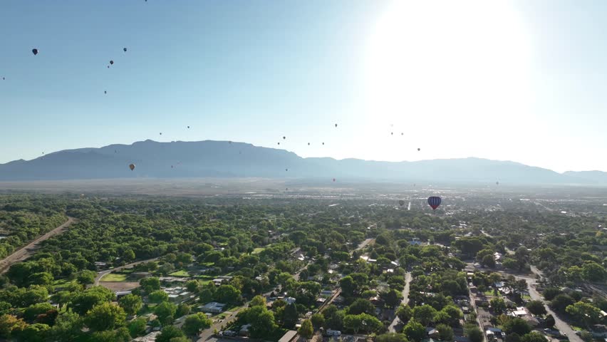 Drone POV of multiple hot air balloons flying over Albuquerque, New Mexico in the early morning near the Sandia Mountains.