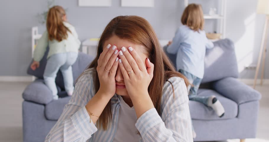 Portrait of tired exhausted mother sitting on floor at home and covering face with hands while noisy kids daughter and son playing pillow fight in background. Problems of parenthood concept. 4k video