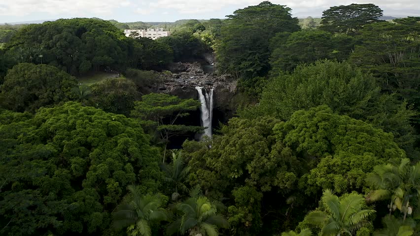 An aerial view of Rainbow Falls on Big Island, Hawaii, beautifully framed by vibrant green rainforest under a bright tropical sky.