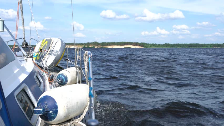 On board a yacht. The sailboat is speeding through the waves. View from the deck. The sandy coastline and forested area are in the background. Fenders on deck.