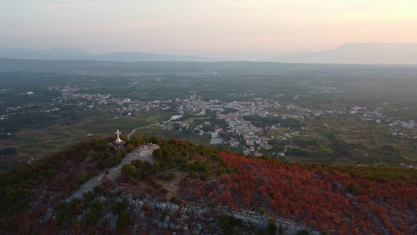 Top of Križevac mountain with white cross. Background of tourist town of Medjugorje, Bosnia and Herzegovina. Aerial view of panorama at sunrise.
