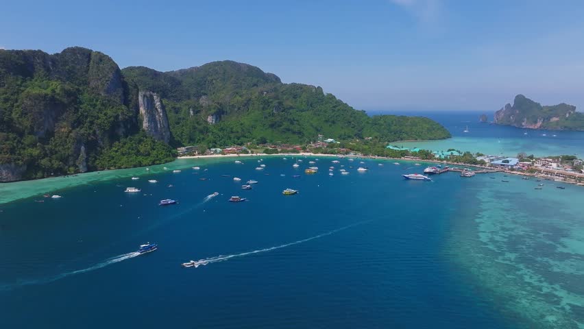 Aerial view of Phi Phi Island in Thailand, featuring turquoise waters, lush green hills, limestone cliffs, boats, and shoreline buildings.