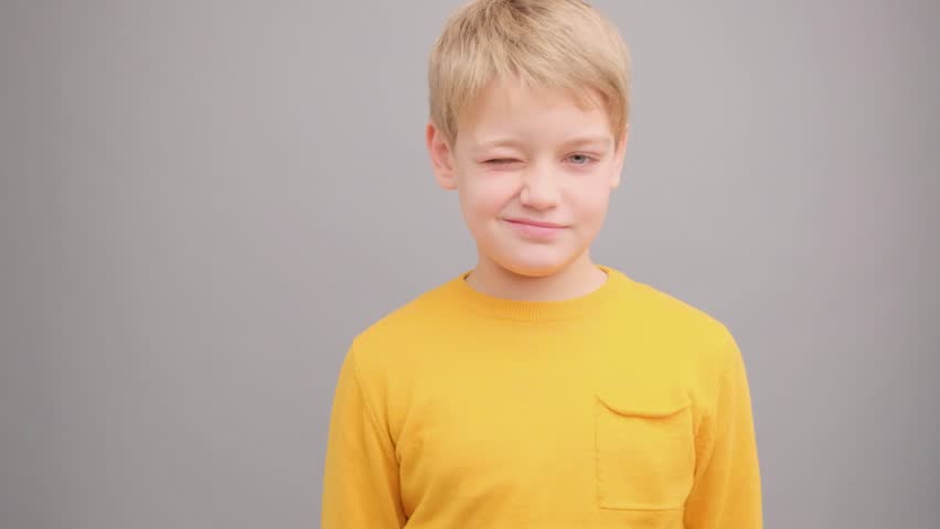 A boy in a yellow T-shirt on a gray background winks and smiles.