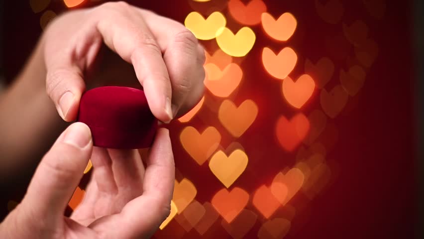 Male hands are opening red box with ring on red background with hearts bokeh. The moment of a Valentine