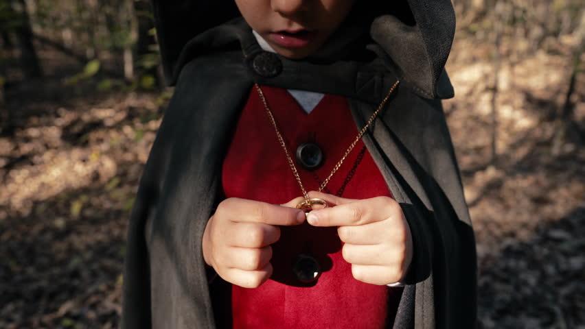 Unrecognizable child cosplayer in hobbit cape costume holding and admiring golden ring. Fantasy journey, halloween, fellowship cosplay