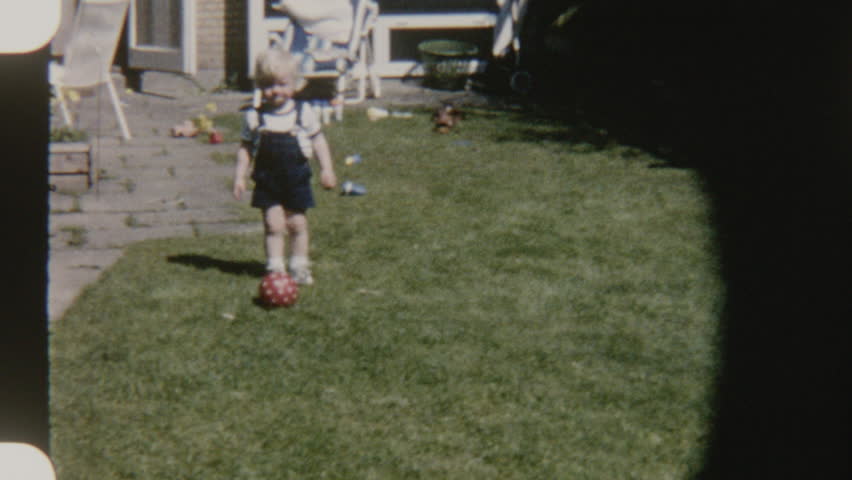 Playful child in a denim jumper kicking a red ball around in a sunny backyard, enjoying outdoor fun on a grassy lawn