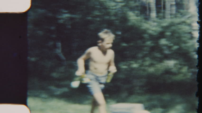 A vintage 8mm film shot captures a boy walking toward the family campsite, holding bottles of refreshments as a thirst quencher while the meal is being prepared in the daylight