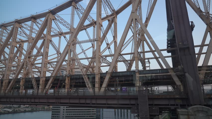 A detailed view of the intricate steel framework of the Queensboro Bridge in New York City. The bridge's structure, captured against the cityscape and river, highlights its industrial design and