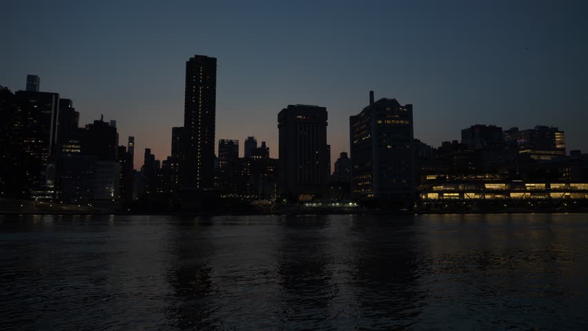 The Manhattan skyline glows with illuminated buildings against the evening sky. The calm East River reflects the city lights, creating a peaceful urban scene.