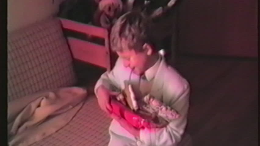 A young boy, surrounded by Christmas presents, sits on a couch intently arranging the items around him in a scene from old home video footage captured on Christmas Eve