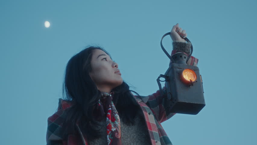 Young Asian woman holding vintage signal lantern with glowing light, gazing upward against clear blue sky at dusk. Low angle view