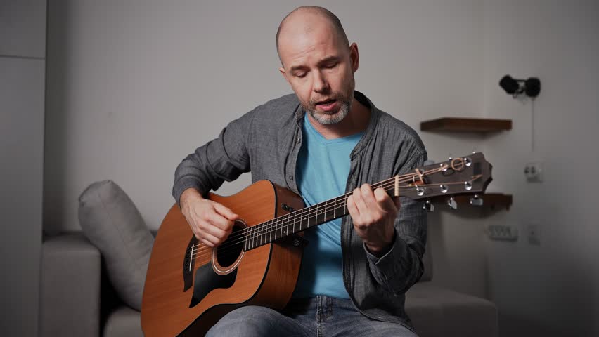 A man passionately plays an acoustic guitar while standing in a simple room. His facial expression reflects the emotional intensity of his music, with a strap securing the guitar around his shoulder.