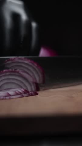 A vertical closeup slow-motion of slicing onion with a kitchen knife on the wooden cutting board