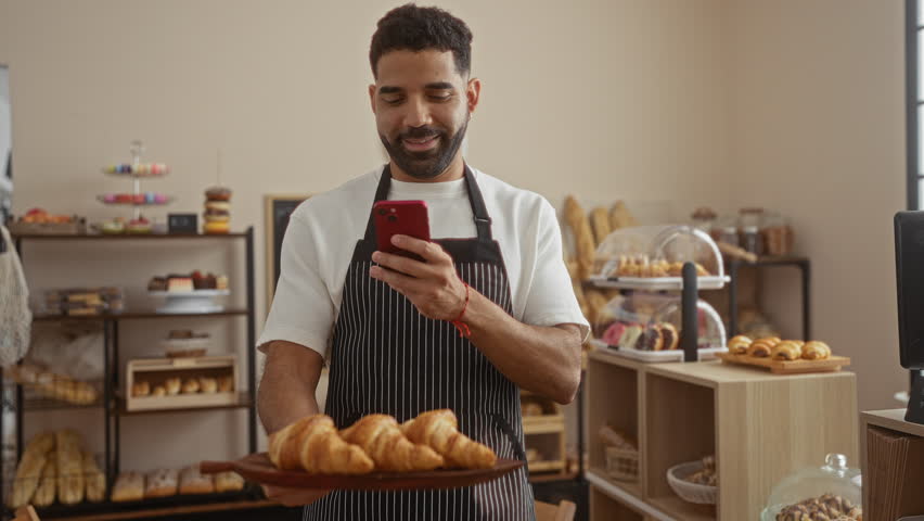 Young man in apron taking a photo of croissants with a mobile phone in a bakery shop interior