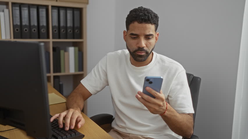 Handsome man working on computer and holding phone in a modern office with shelves of binders in the background