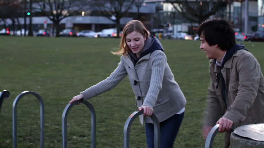 Attractive young couple play by swinging on bike racks. Medium long shot at dusk