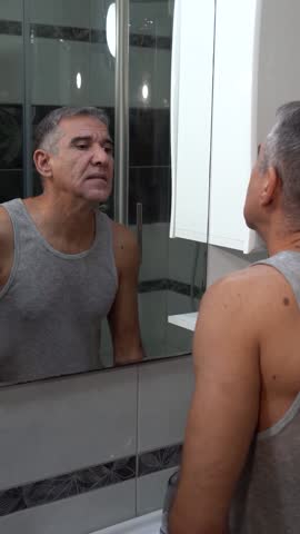 Man Finishing Shaving Routine in Front of Mirror. Medium shot of a middle-aged man checking skin smoothness and drying his face after shaving. Perfect for skincare routines and wellness content.